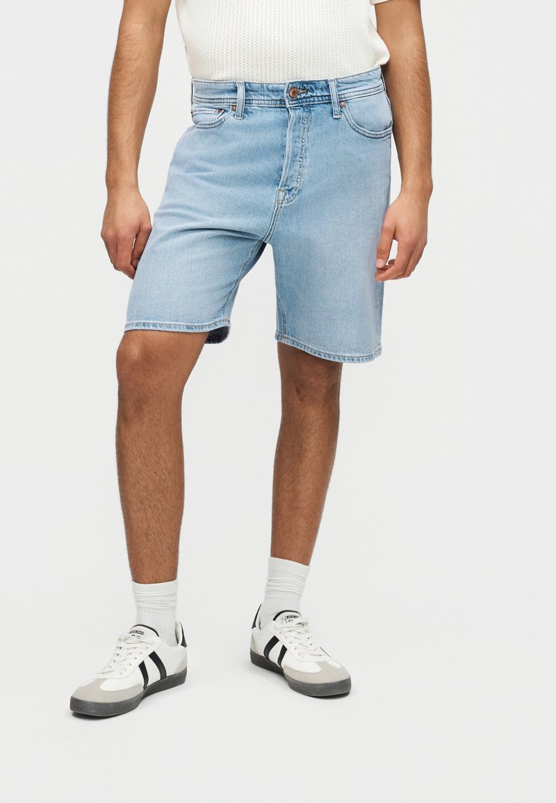Person som har på seg lyseblå denimshorts, hvite sokker, hvite og svarte joggesko, og en hvit teksturert kortermet topp, stående mot en ensfarget bakgrunn.