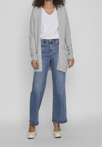 Grå stickad kofta, vit t-shirt med v-ringning, ljusblå vida jeans och krämfärgade plattformsskor. Anmärkningsvärda detaljer inkluderar fickor och ribbade ärmslut.