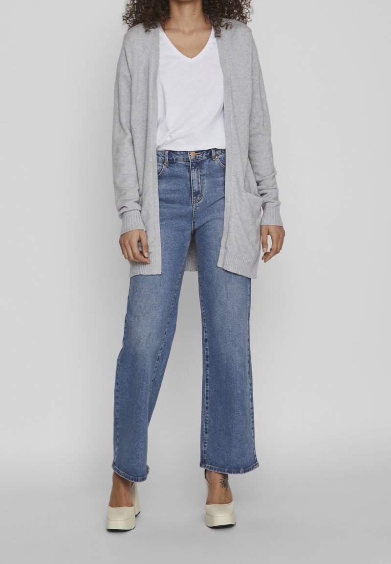 Grå stickad kofta, vit t-shirt med v-ringning, ljusblå vida jeans och krämfärgade plattformsskor. Anmärkningsvärda detaljer inkluderar fickor och ribbade ärmslut.