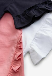 Trois tops à manches courtes : navy, rose et blanc. Chacun présente des détails de bas de vêtement volants, un tissu en coton doux et une coupe ajustée.