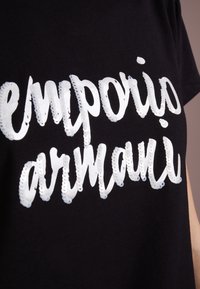Svart t-shirt med en stor, glänsande vit broderad logotyp som säger "emporio armani" i en flödande skriftsstil med texturerad yta.