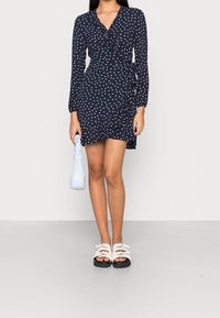 Eine Frau in einem marineblauen Pünktchen-Wickelkleid, die eine hellblaue Handtasche hält, kombiniert mit weißen Sandalen mit offenen Zehen und dicken schwarzen Sohlen.