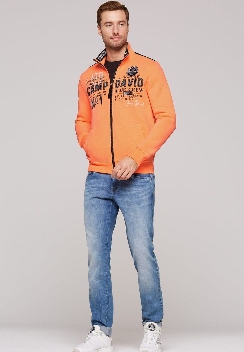 Felpa arancione con zip, decorata con testo stampato e dettagli grafici, abbinata a jeans azzurri chiari e sneaker bianche. Tessuto liscio, vestibilità casual.