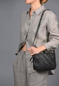 Bolso de mano de cuero trenzado negro con forma rectangular y una sola correa ajustable, sostenido por una persona que lleva un conjunto de lino gris.