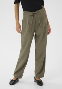 Pantalon taille haute vert olive à jambes larges, avec un devant plissé et une taille à cordon, associé à des chaussures plates noires.