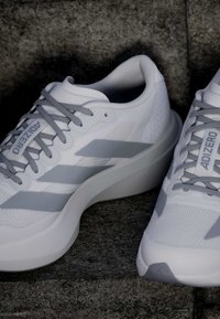 Λευκά και γκρι παπούτσια για τρέξιμο Adidas Adizero με κορδόνια, τοποθετημένα σε σκοτεινή πέτρινη επιφάνεια.