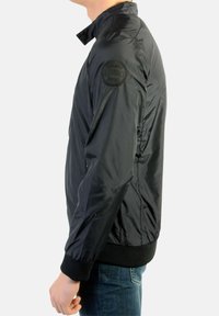 Sort polstret bomberjakke med høj krave, der har et logopatch på venstre skulder samt elastiske manchetter og kant. Bær med blå jeans.