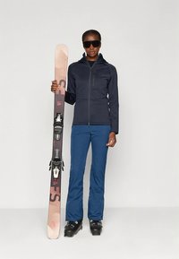 Zilā kalnu slēpošanas jaka, zilas kalnu slēpošanas bikses un melni kalnu slēpošanas zābaki. Tur rozā slēpi ar melniem stiprinājumiem. Aizsargbrilles papildina tēlu.