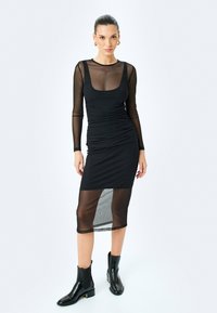 Czarna sukienka z długimi przezroczystymi rękawami, z marszczeniem i długości midi, z przezroczystym panelem u dołu. Noszona z czarnymi botkami za kostkę.