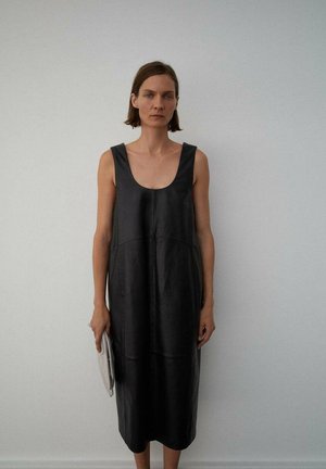 Vestido sin mangas de cuero negro con escote redondo, corte relajado y textura suave. La modelo sostiene un bolso de mano gris claro.