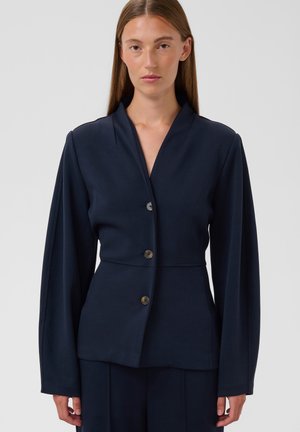 Femme aux cheveux lisses portant un blazer bleu marine foncé ajusté à trois boutons et un pantalon assorti sur un fond uni.