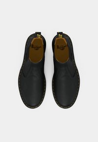 Een paar zwarte leren instapschoenen van Dr. Martens met gele stiksels en merkpulltabs, bekeken vanuit boven op een witte achtergrond.