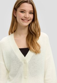 Kremowy sweter kardigan z dekoltem w serek, prążkowanym kołnierzem i zapięciem na guziki. Miękka tekstura i luźny fason. Nosi się go na czarną bluzkę.