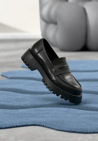 Schwarzer Leder-Slipper mit dicker, robuster Sohle und überlagerter Front-Riemendetail, auf einem strukturierten blauen Teppich platziert.
