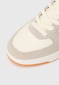 Wildleder- und synthetischer Sneaker mit einem cremefarbenen Obermaterial und hellgrauen Akzenten, runden Schnürsenkeln und einer strukturierten Gummisohle in Kautschuk.