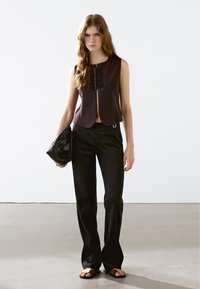 Mouwloze, donker bordeaux top met een sluiting aan de voorkant, gestructureerd paneel, gecombineerd met zwarte flared broeken en een geweven zwart clutch. Bruine sandalen.