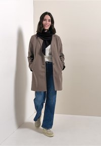 Brauner Wollmischmantel mit lockerer Passform, kombiniert mit hellblauen, weit geschnittenen Jeans und einem schwarzen Schal, vor einem neutralen Hintergrund.