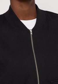 Czarna kurtka bomberka zapinana na zamek, z prążkowanym kołnierzem i gładką fakturą. Posiada boczną kieszeń i minimalistyczny design.