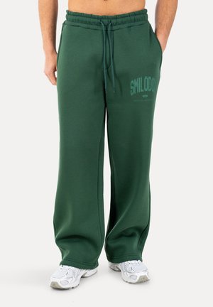 Mężczyzna w luźnych ciemnozielonych spodniach dresowych z logo „SMILODOX” i białych trampkach, stojący na białym tle.