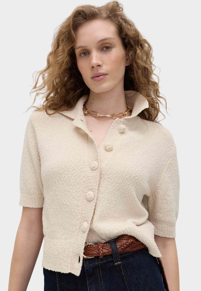 Femme aux cheveux bouclés portant un cardigan beige à manches courtes avec des boutons recouverts de tissu, un collier chaîne en or massif et un jean en denim foncé.