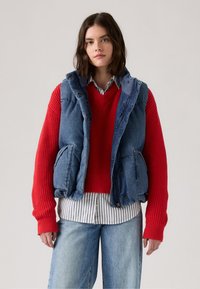 Denim vest met een capuchon, twee voorzakken; gelaagd over een rode geribde trui en een gestreept shirt, gecombineerd met lichtblauwe jeans.