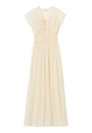 Mujer con vestido largo sin mangas de color crema, tacones negros con tiras y una gorra de béisbol azul marino, de pie frente a un fondo claro y liso.