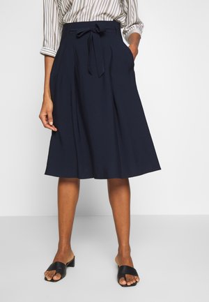 Femme portant une jupe bleu marine longueur genou avec une ceinture nouée, un chemisier rayé et des sandales à talons ouvertes noires.