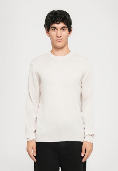 Jeune homme aux cheveux bouclés portant un pull à col rond beige clair et un pantalon noir, se tenant devant un fond blanc uni.