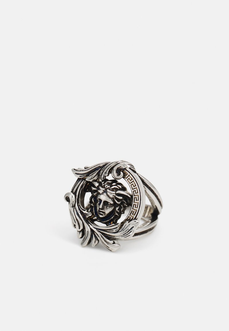 Anillo de plata con un diseño circular que presenta una cara detallada en el centro, rodeada de hojas ornamentadas y acentos con patrones griegos.