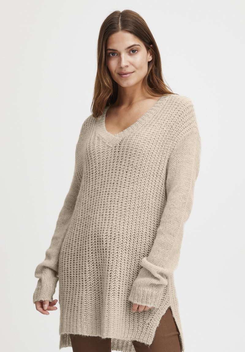 Beżowy sweter tunika z dzianiny, z dekoltem w serek, długimi rękawami, prążkowanymi mankietami oraz bocznymi rozcięciami. Teksturowany materiał o luźnym kroju.