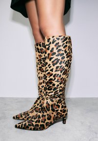 Des bottes aux genoux en imprimé léopard fabriquées en tissu texturé, présentant un bout pointu et un talon mince et structuré. La palette de couleurs comprend des tons beige, noir et marron.
