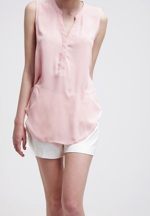 Blusa sin mangas de color rosa claro con escote en V y pantalones cortos blancos, llevada por una persona de pie contra un fondo liso.