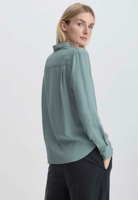 Camicia lunga color turchese chiaro realizzata in tessuto morbido con una leggera lucentezza, dotata di colletto con bottoni e orlo arrotondato.