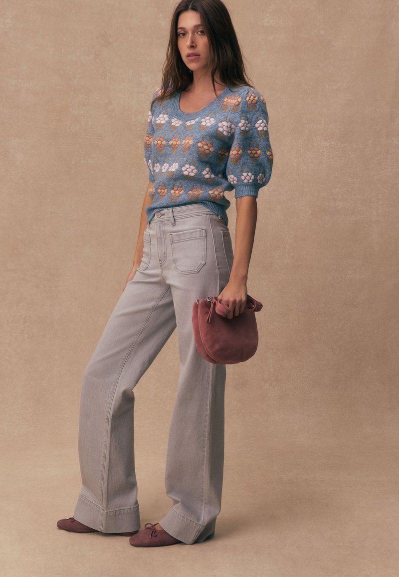 Femme portant un pull bleu à manches bouffantes à fleurs, un pantalon large gris clair, et tenant un petit sac à main mauve.