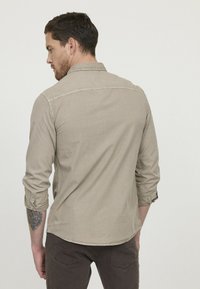 Chemise beige clair à manches longues, avec des poignets boutonnés, des coutures discrètes sur les épaules, coupe décontractée, texture douce. Portée avec un pantalon sombre.