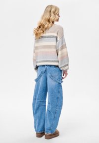Sweter dziany w beżowe, niebieskie i różowe paski, zestawiony z dużymi jasnoniebieskimi dżinsami z unikalnymi wycięciami i paskami. Brązowe obuwie.