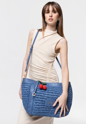 Donna con vestito beige senza maniche che porta una grande borsa intrecciata blu con decorazione di rosa rossa sulla spalla su sfondo bianco.