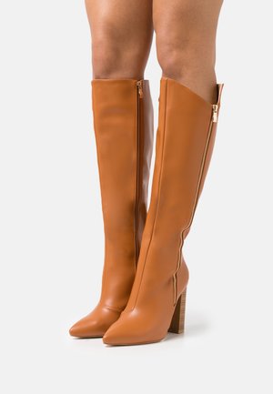 Bottes en cuir camel montant jusqu'aux genoux avec fermetures éclair latérales, bouts pointus et talons carrés portées sur des jambes nues sur un fond uni.
