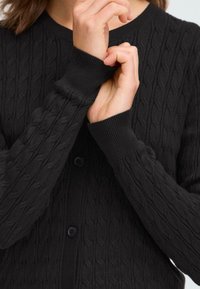 Cardigan noir en tricot à torsades avec des poignets côtelés et une fermeture à boutons. La texture est douce avec un motif en relief détaillé sur l'ensemble.