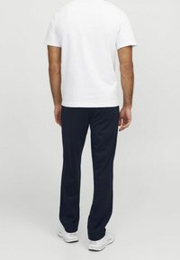 Uomo con camicia bianca a maniche corte, pantaloni blu navy e sneakers bianche, che sta in piedi di spalle contro uno sfondo chiaro e semplice.