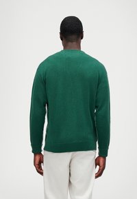 Πράσινη πλεκτή ζακέτα με κλασικό λαιμόκοψη, ριμπ μανσέτες και στρίφωμα, που αναδεικνύει ένα υφασμάτινο σχέδιο. Φοριέται με ανοιχτόχρωμο παντελόνι.