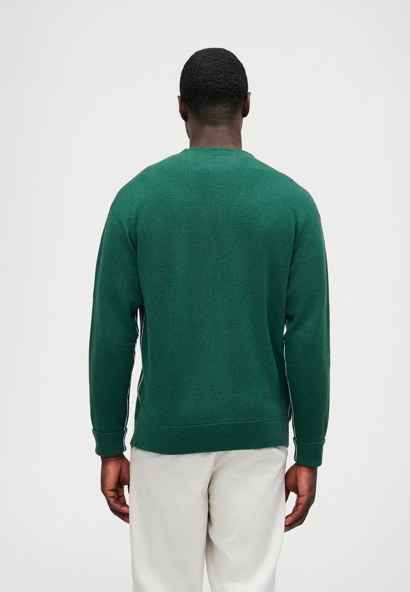 Suéter de punto verde con cuello redondo, puños y dobladillo acanalados, que muestra un tejido texturizado. Usado con pantalones de color claro.