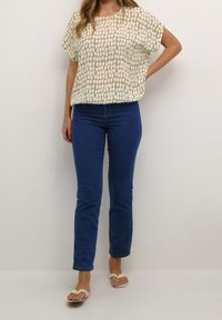 Cremefarbige Bluse mit Muster und kurzen Ärmeln, kombiniert mit blauen Jeans. Die Bluse hat ein strukturiertes Gitterdesign. Leichte, lässige Schuhe runden den Look ab.