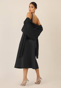 Vestido de satén negro con tirantes ajustables, de longitud a media pantorrilla, con una chalina de capas que cae sobre los hombros; sandalias de tacón alto plateadas.