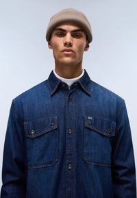 Camisa de mezclilla en azul oscuro con dos bolsillos en el pecho, cierre de botones y costuras en contraste. El modelo lleva un gorro de punto de color marrón claro.