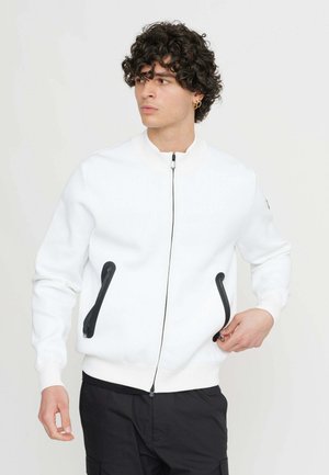 Giacca bomber bianca con tasche a zip nere, colletto e polsini a coste, e una texture liscia. Presenta una zip frontale e un design minimalista.