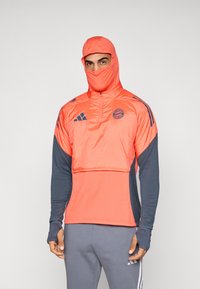 Άνδρας που φορά αθλητική ένδυση σε κοραλλί και γκρι απόχρωση με καλυμμένο πρόσωπο από κουκούλα, ορατά τα λογότυπα της FC Bayern Munich και της Adidas στο επάνω μέρος του στήθους.