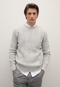 Grauer gestrickter Pullover mit einem strukturierten Zopfmuster, ausgestattet mit einem Rundhalsausschnitt und gerippten Bündchen, über einem weißen Hemd mit Kragen getragen.