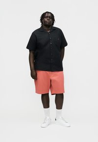 Mężczyzna o ciemnej karnacji i splecionych włosach stoi, ubrany w czarną koszulę na krótki rękaw z guzikami, koralowe szorty, białe skarpetki i białe trampki na białym tle.