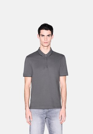 Giovane uomo che indossa una polo a maniche corte grigio scuro tinta unita e jeans grigio chiaro, in piedi contro uno sfondo bianco.